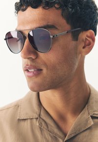 Naočale za sunce s tamnim gradijentnim lećama, metalnim okvirom i tankim kracima. Prikaz izbliza koji pokazuje crte lica i svijetlosmeđu košulju.