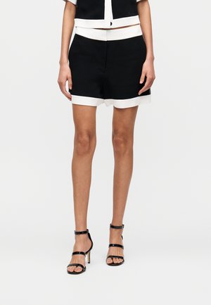 Svarte shorts med en hvit kant nederst, med en avslappet passform. Kombinert med svarte høyhælte sandaler med stropper. Glatt stofftekstur.