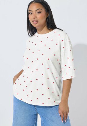 Ung kvinde med lige sort hår iført en hvid oversized T-shirt med små røde hjerter og lyseblå jeans, stående mod en ensfarvet baggrund.