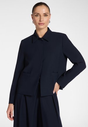 Vrouw draagt een op maat gemaakte marineblauwe blazer met kraag en bijpassende wijde broekspijpen, poseert met één hand op haar heup tegen een witte achtergrond.