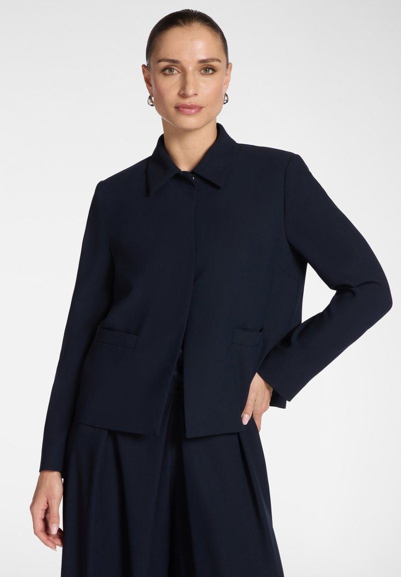 Mujer con una blazer azul marino a medida con cuello y pantalones de pierna ancha a juego, posando con una mano en la cadera sobre un fondo blanco.