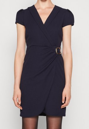 Frau trägt ein kurzärmeliges, marineblaues Wickelkleid mit V-Ausschnitt und goldener Schnallendetail am Taillenbund, kombiniert mit schwarzen, transparenten Strumpfhosen.