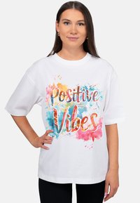 Wit oversized T-shirt met de tekst "Positive Vibes" in kleurrijk handschrift, gedrukt op een achtergrond van getextureerde aquarelspatten.