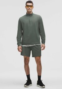 Zelený ľahký pulóver s polovičným zipsom, skombinovaný s odpovedajúcimi šortkami. Tkanina je štruktúrovaná, s atletickými topánkami dotvárajúcimi outfit.