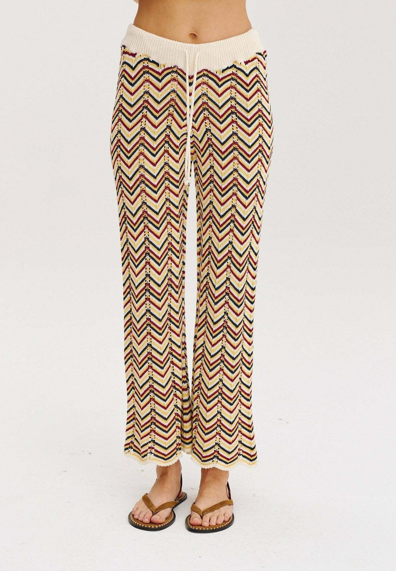 Dzianinowe spodnie o szerokich nogawkach, z beżową bazą ozdobioną wielokolorowymi wzorami w zygzak. Elastyczny pasek w talii z detalem sznurka. Płaskie sandały.
