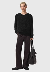 Zwarte gebreide trui met ribbeltextuur en oversized pasvorm, gecombineerd met losse donkere broeken en een slungelige zwarte tas. Simpele, monochrome uitstraling.