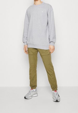 Persona che indossa una felpa grigio chiaro, pantaloni cargo verde oliva con tasche e sneakers grigio chiaro con loghi New Balance rossi.