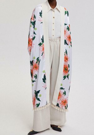 Persoon die beige wijde broek draagt, een crèmekleurig hemd met knopen en een lange witte kimono-jas met oranje bloemen en groene bladeren.