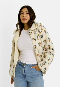 Veste zippée en velours crème avec un motif floral rose, marron et vert. Elle dispose d'une capuche et de poignets côtelés ; portée avec un débardeur blanc et un jean.