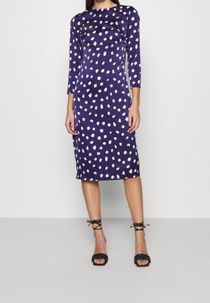 Kobieta w granatowej sukience do kolan w białe kropki z rękawami do łokcia, w połączeniu z czarnymi sandałami na obcasie z paskami.