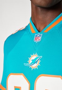 Ένα τιρκουάζ NFL τζέρσεϊ των Miami Dolphins με πορτοκαλί λεπτομέρειες. Διαθέτει το λογότυπο ενός δελφινιού, αριθμό 88 και ραμμένο έμβλημα NFL στον γιακά.
