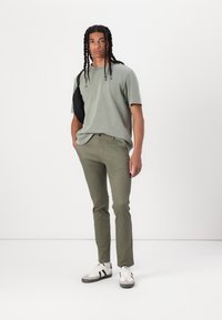 T-shirt olive à manches courtes, pantalon olive slim-fit, sac bandoulière noir, baskets blanches avec rayures noires, debout contre un fond blanc.