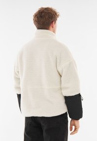 Hvid fleece-pullover med høj krave, sorte ærmer i kontrast og en afslappet pasform. Blød tekstur med en sømdetalje på tværs af ryggen.
