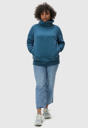 Un sweatshirt en bleu-vert avec un col cheminée, orné de pois blancs et de deux boutons marron. Accompagné d'un jean court bleu clair et de baskets grises.