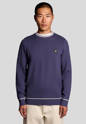 Man draagt een marineblauwe trui met witte bies en een klein geel vogellogo op de borst, staand tegen een effen lichtgrijze achtergrond.