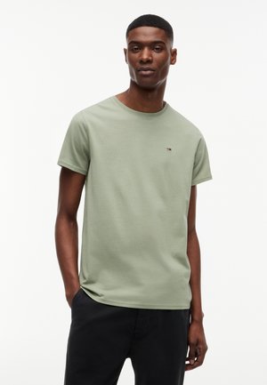 Bărbat cu păr scurt, purtând un tricou verde deschis cu mânecă scurtă și un mic logo pe piept, și pantaloni negri, stând cu o mână în buzunar.