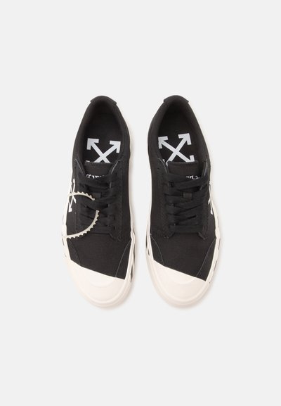 Ζευγάρι μαύρα πάνινα sneakers με λευκές λαστιχένιες μύτες και λογότυπα βελών στις εσωτερικές σόλες και τις πλευρές, εμφανίζονται από πάνω σε λευκό φόντο.