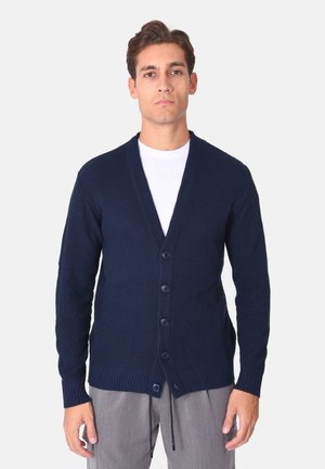 Cardigan a maglia blu navy con scollo a V, chiusura con cinque bottoni, trama a coste e maniche lunghe, indossato sopra una camicia bianca.