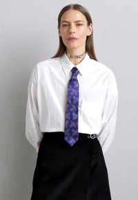 White oversized button-up shirt with a pocket, paired with a dark corduroy skirt. Features a blue tie with a pink pattern and a metallic choker.