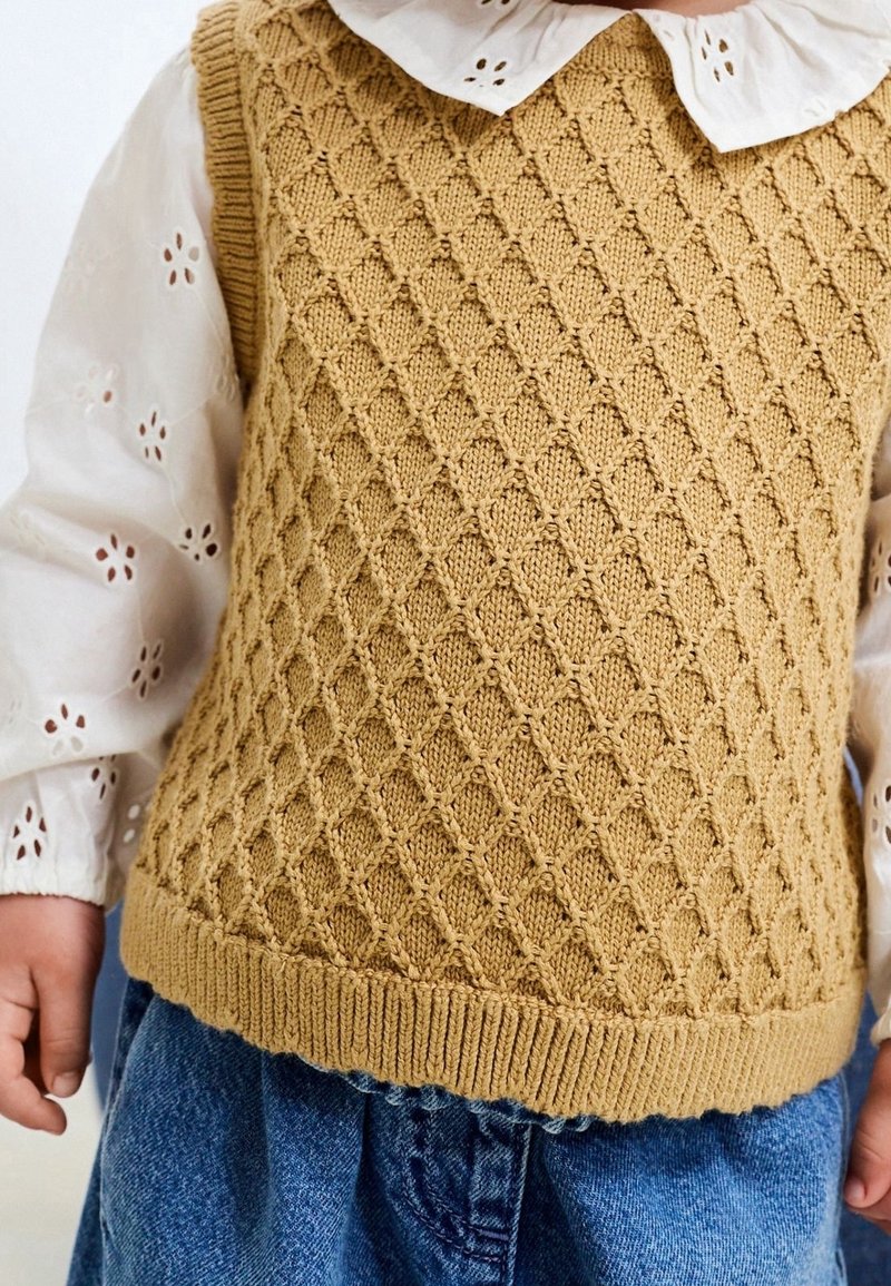 Enfant portant un gilet tricoté beige texturé par-dessus une blouse blanche à œillets avec un col à volants et un jean en denim bleu.