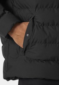 Chaqueta de plumas negra con un exterior texturizado, costuras horizontales prominentes y un bolsillo lateral con cierre a presión.