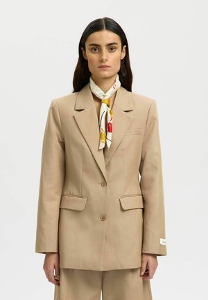 Kvinne med langt mørkt hår iført en beige skreddersydd blazer og matchende bukser, tilbehør som et hvitt skjerf med gule og røde mønstre.