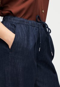 Άτομο που φοράει σκούρο μπλε τζιν παντελόνι με ελαστική μέση και κορδόνι, χέρι στην τσέπη, σε συνδυασμό με καφέ πουκάμισο με κουμπιά.