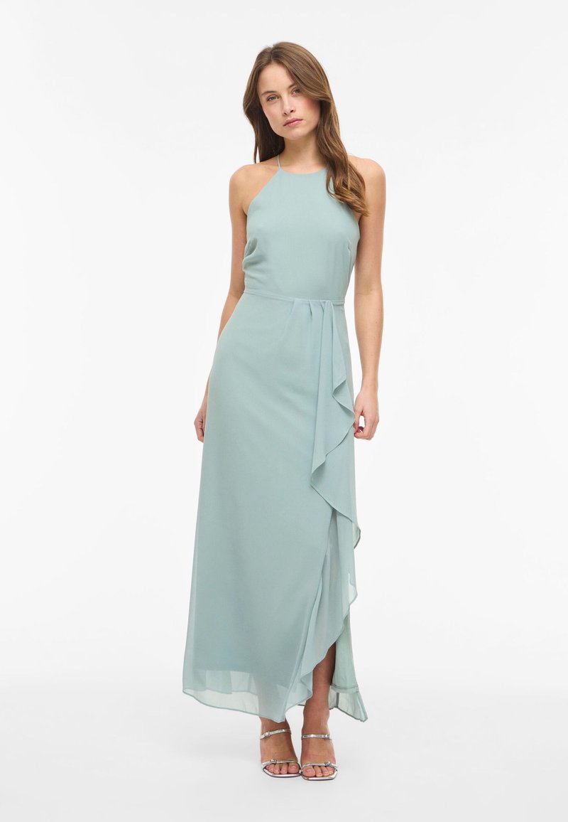 Femme portant une robe maxi bleu clair sans manches à col licou avec une fente latérale à volants et des talons ouverts argentés, debout devant un fond blanc.