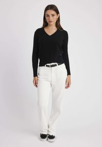 Pull noir à col en V avec des manches longues, associé à un pantalon blanc à coupe droite et une ceinture noire. Le modèle porte des baskets noir et blanc.