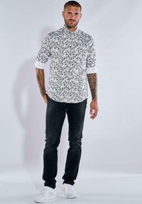 Gemustertes Langarmhemd in Schwarz und Weiß mit Kragen, hochgerollten Ärmeln, schwarzen Slim-Fit-Jeans und weißen Sneakern.