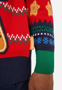 Punane kootud kampsun, millel on pühade mustrid, sealhulgas tähed ja lumememmed, tumesiniste detailidega ning rohelise soonikkoega mansetiga. Silt on nähtaval.