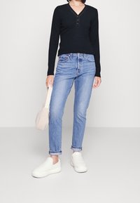 Czarny długi rękaw top z dekoltem w serek i trzema guzikami, zestawiony z jasnoniebieskimi dżinsami i białymi teksturowanymi butami wsuwanymi. Niesie małą beżową torbę.