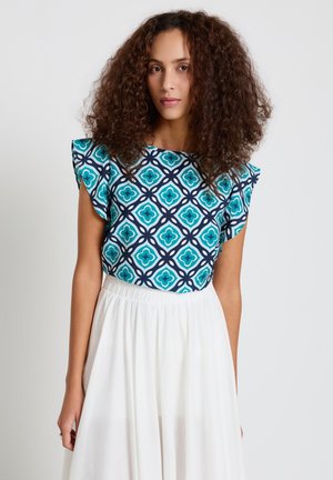 Frau mit lockigem Haar, die ein blau gemustertes, ärmelloses Oberteil und einen hoch taillierten weißen Rock vor einem einfarbigen weißen Hintergrund trägt.