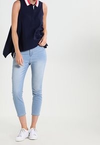 Top senza maniche di colore blu navy con colletto rosso e bianco, abbinato a jeans slim-fit blu chiaro e sneakers bianche. Tessuto morbido e design casual.