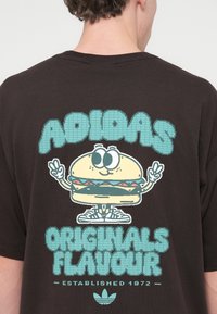 Μαύρη μπλούζα με στάμπα καρτούν χαρακτήρα burger με μεγάλα μάτια, χέρια και σήμα ειρήνης, μαζί με το κείμενο "ADIDAS ORIGINALS FLAVOUR".