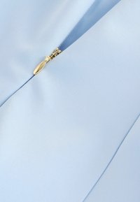 Gaiši zils audums ar daļēji atvērtu zelta rāvējslēdzēju, redzamas auduma struktūras un šūšanas detaļas.