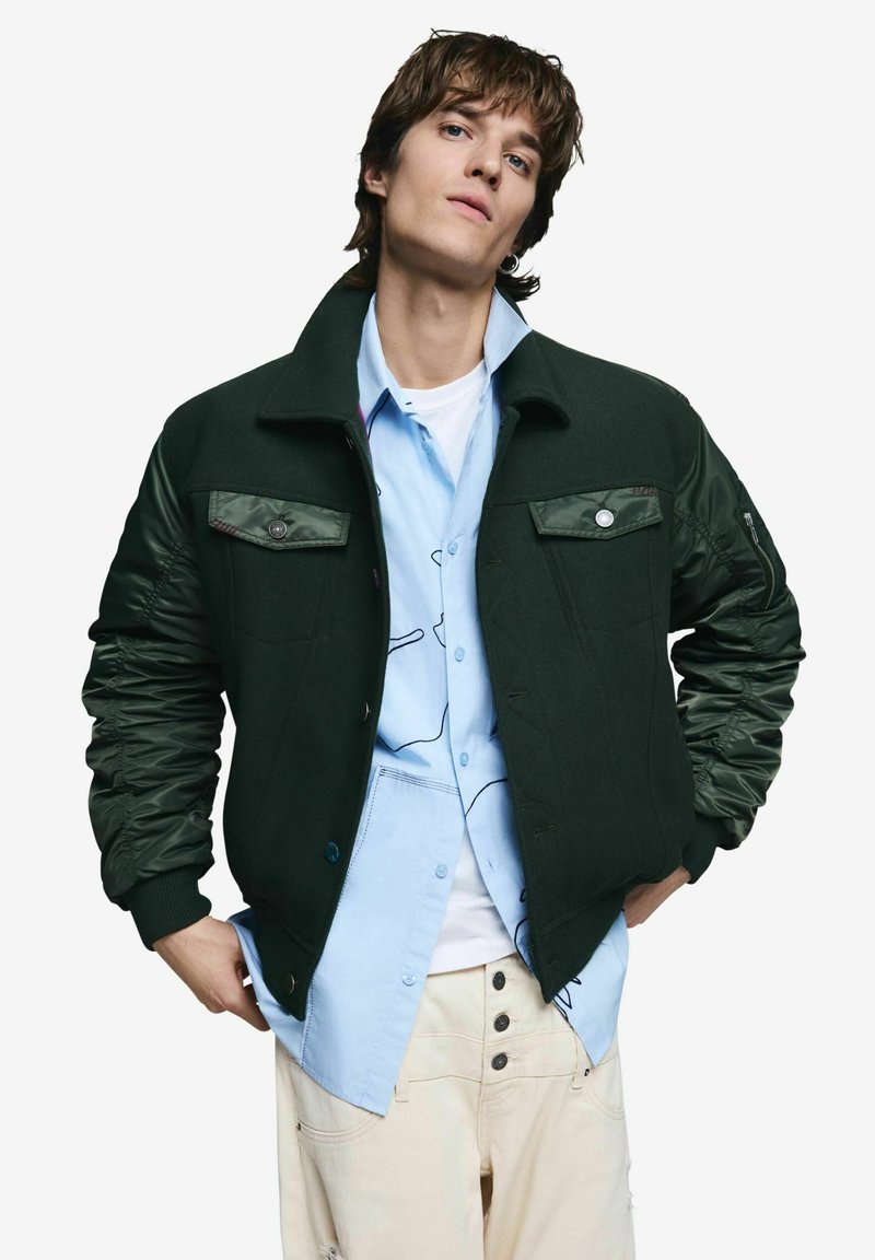 Ung mand iført en mørkegrøn bomberjakke over en lyseblå skjorte med abstrakte mønstre og cremefarvede bukser, poserer med hænderne i lommerne.