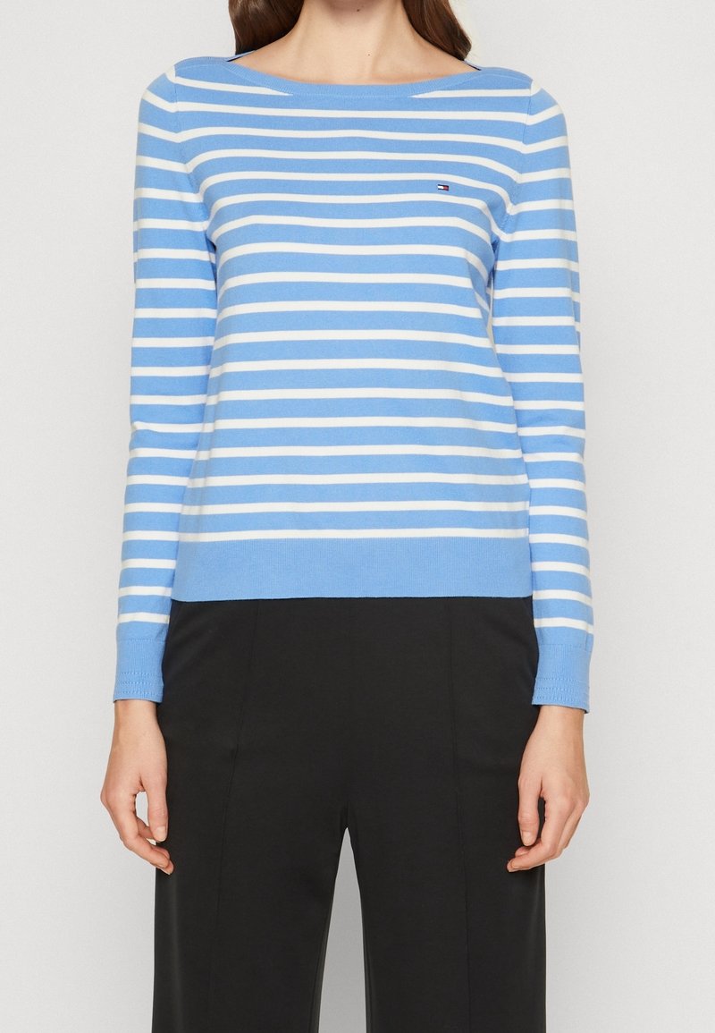 Kobieta ubrana w jasnoniebieską i białą poziomą paskowaną bluzkę z długim rękawem oraz czarne spodnie z wysokim stanem, stojąca na tle jednolitym.