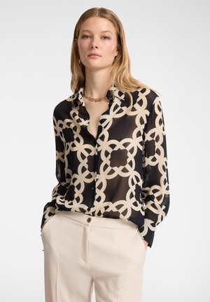 Blusa nera e crema con motivo, caratterizzata da una texture trasparente, con colletto a bottone e maniche lunghe, abbinata a pantaloni beige chiaro.