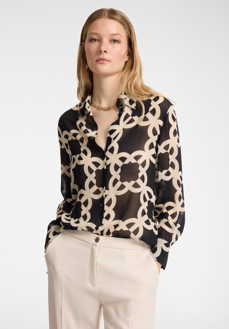 Blusa nera e crema con motivo, caratterizzata da una texture trasparente, con colletto a bottone e maniche lunghe, abbinata a pantaloni beige chiaro.