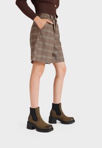 Donna che indossa pantaloni corti a quadri marroni fino al ginocchio, un maglione marrone scuro e stivali alla caviglia con tacco blocco verde oliva e lati elastici neri.