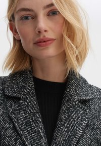 Black-and-white houndstooth coat with wide lapels, textured fabric, layered over a black ribbed sweater. Close-up of face with light hair.