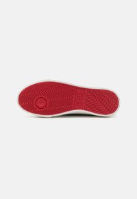 Rubber sneaker outsole featuring a red color with zigzag tread pattern and circular logo. White edge and textured surface provide grip.