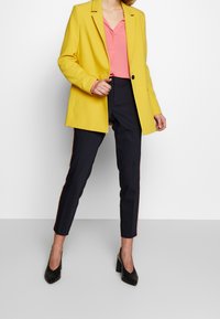 Yellow blazer with a single button, pink blouse underneath, paired with navy pants featuring red side stripes. Black high-heeled shoes.