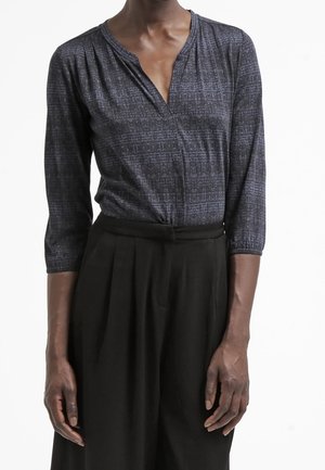 Personne portant une chemise à manches longues à motifs sombres, rentrée dans un pantalon noir plissé taille haute, sur un fond blanc.