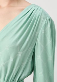 Bluză verde cu decolteu drapat, având un model geometric cu frânghii împletite și logo, realizată dintr-un material ușor.