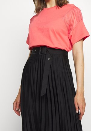 Kobieta ubrana w jasnoróżową koszulkę Adidas, wpuszczoną w czarną plisowaną spódnicę z wysokim stanem i dopasowanym paskiem, stojąca na jednolitym tle.