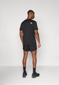 Schwarzes T-Shirt mit kurzen Ärmeln und einem kleinen weißen Logo auf dem Rücken, kombiniert mit schwarzen Shorts und dunklen Sportschuhen. Schlichtes und funktionales Design.