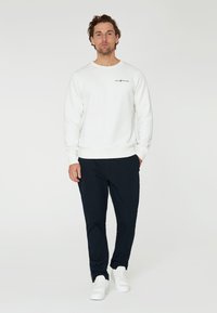 Sudadera blanca con cuello redondo y logotipo negro, combinada con pantalones navy ajustados y zapatillas blancas, sobre un fondo neutro.