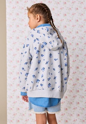 Młoda dziewczyna z warkoczami, ubrana w szarą bluzę z kapturem z niebieskimi kwiatowymi wzorami, na niebieskiej koszuli i dżinsowych szortach.